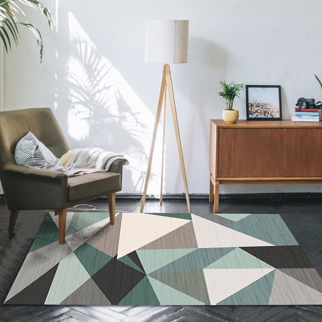 la alfombra de la sala de estar es geométricamente buena para uso doméstico, el dormitorio está cubierto con tapetes y la alfombra de la mesita de noche se usa como tapete Alfombra geométrica multicolor para sala y dormitorio; vista frontal con patrón de triángulos y vetas tipo madera en tonos verde, gris y beige; colocada sobre piso oscuro frente a sillón gris y lámpara de trípode, con aparador de madera y planta al fondo. Adecuada como tapete junto a la mesita de noche.