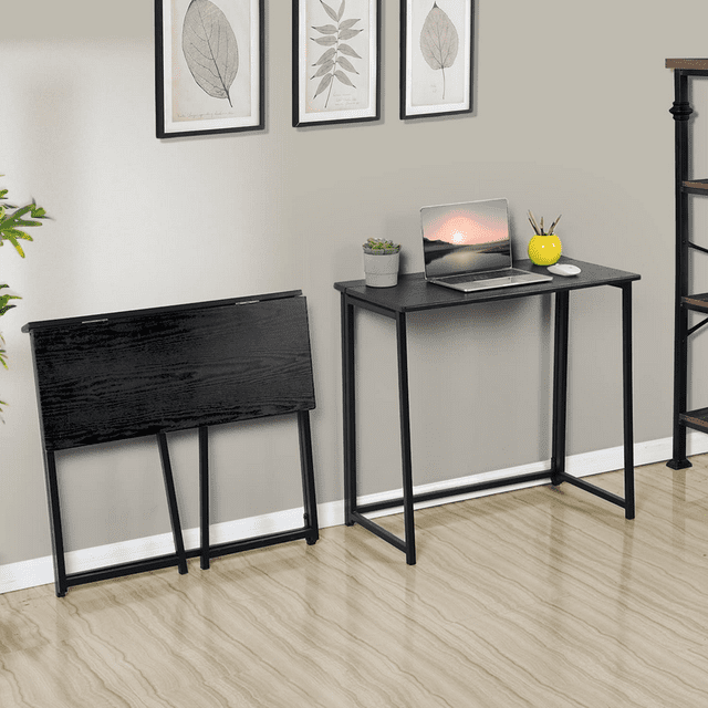 escritorio homemake furniture plegable rectangular industrial clásico negro en posición frontal; mesa compacta con superficie lisa de madera de ingeniería negra y estructura tubular de metal negro plegable, una unidad montada con laptop y maceta y otra guardada plegada contra la pared; ideal para espacios reducidos y uso en dormitorio u oficina, a menudo usado como estación de estudio portátil para regreso a clases.