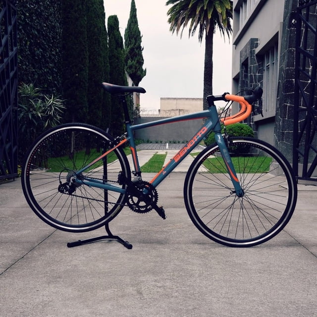 bicicleta benotto ruta 590 r700 14v aluminio palancas duales gris oscuro 51 — vista lateral frontal-izquierda de ruta con cuadro de aluminio 6061 gris oscuro y logo Benotto naranja, manubrio de carretera con cinta naranja, palancas duales Microshift para freno y cambio, desviador y cambio trasero Microshift, transmisión 14 velocidades con platos 48x38 y eje de centro sellado, llantas 700x23c y sillín ergonómico; ubicada en patio exterior pavimentado.