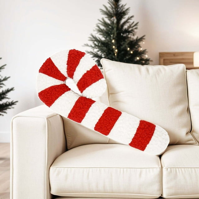 cojín navideño con forma de bastón de caramelo | cojín decorativo rojo y blanco con forma de menta, vista 3/4 apoyado en sofá beige; felpa blanca con franjas rojas texturadas y curva superior en bastón, costuras discretas y aspecto mullido, fondo con árbol de Navidad desenfocado. Adecuado como adorno estacional para sala, dormitorio u oficina.