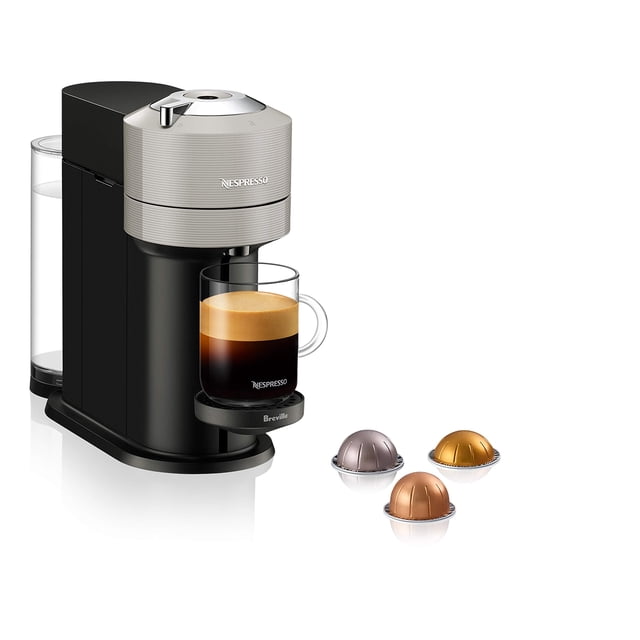 cafetera y cafetera espresso nespresso vertuo next breville negra, vista frontal 3/4 con tanque de agua extraíble transparente de 37 oz atrás, cabezal superior plateado con logo Nespresso, bandeja de goteo ajustable con taza de vidrio llena de espresso con crema y tres cápsulas Nespresso en primer plano; especificación: 40% plástico reciclado; prepara café o espresso de un solo servicio con un botón, adecuada también para uso rápido en oficinas pequeñas.