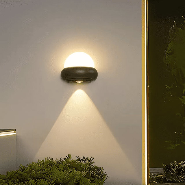 lámpara de pared skandiglow circular iluminacion exterior negro 7w 12w vista frontal centrada de lámpara LED de pared exterior en negro; cúpula translúcida que emite luz y base oblonga de PC y aluminio negra que proyecta un cono de luz hacia abajo. Para jardín, patio o pasillo; luz blanco frío o blanco cálido, IP65, 7W/12W; también adecuada para iluminación decorativa de entradas y terrazas.