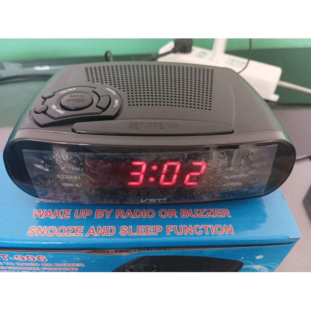 radio despertador controlado por reloj con canal AM y FM Radio despertador LED con función de repetición Reloj LED rojo ENTASSER, vista frontal centrada de un despertador compacto con pantalla LED roja que muestra 3:02, botones de radio/encendido y sintonizador en la parte superior, rejilla de altavoz y superficie plástica reflectante, apoyado sobre una caja promocional; a menudo usado como despertador infantil por su tamaño compacto y función de repetición.