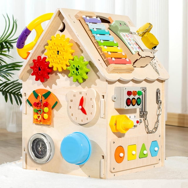 actividad material de tablero ocupado habilidades básicas habilidad de aprendizaje juguete accesorios de bricolaje juguetes educativos juguete de estilo d cuticat tablero de niños ocupados Estilo D: vista frontal 3/4 de una casita de actividades de madera con asa amarilla y techo inclinado; xilófono arcoíris en el techo, engranajes rojos/amarillo/verde giratorios, reloj de madera con manecillas rojas, cadena metálica con pestillo, botón giratorio amarillo, fila de botones de colores, brújula integrada y pomo azul circular, y panel con encaje de formas geométricas (círculo, cuadrado, triángulo). Fondo interior desenfocado. Adecuado para entrenamiento de motricidad fina y estimulación del pensamiento creativo.
