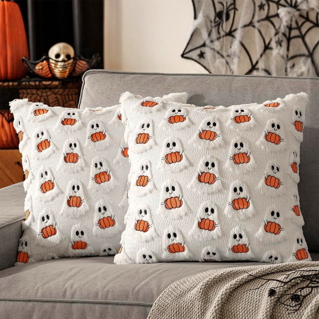 funda de almohada de fantasma de halloween, cojín decorativo para sala de estar o sofá multicolor: dos fundas cuadradas 45x45 cm en felpa blanca con estampado repetido de pequeños fantasmas sosteniendo calabazas naranjas, textura suave visible y cierre con cremallera oculta; colocadas sobre un sofá gris con decoración de Halloween al fondo.