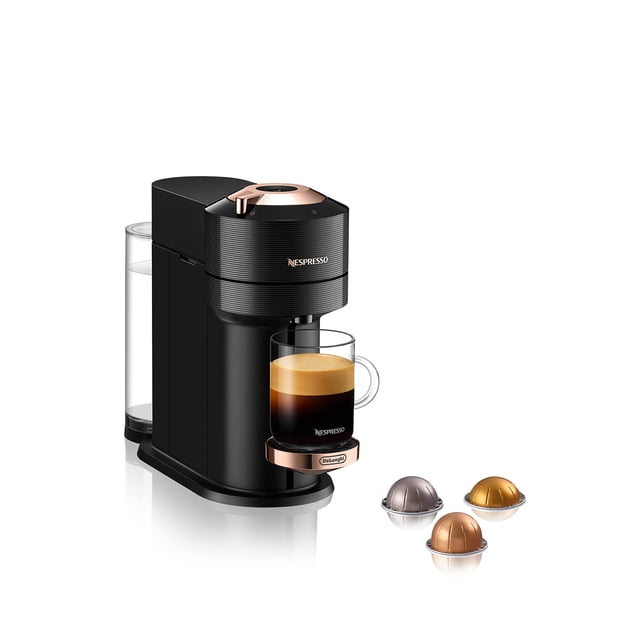 máquina de café y espresso nespresso vertuo next de'longhi negro con oro rosa, vista 3/4 frontal con depósito de agua trasero transparente, bandeja recoge gotas y taza transparente con espresso y crema en la salida; panel superior y base en acabado oro rosa y tres cápsulas Vertuo delante que muestran tamaños variados; suele usarse también para preparar cafés para llevar en tazas térmicas.