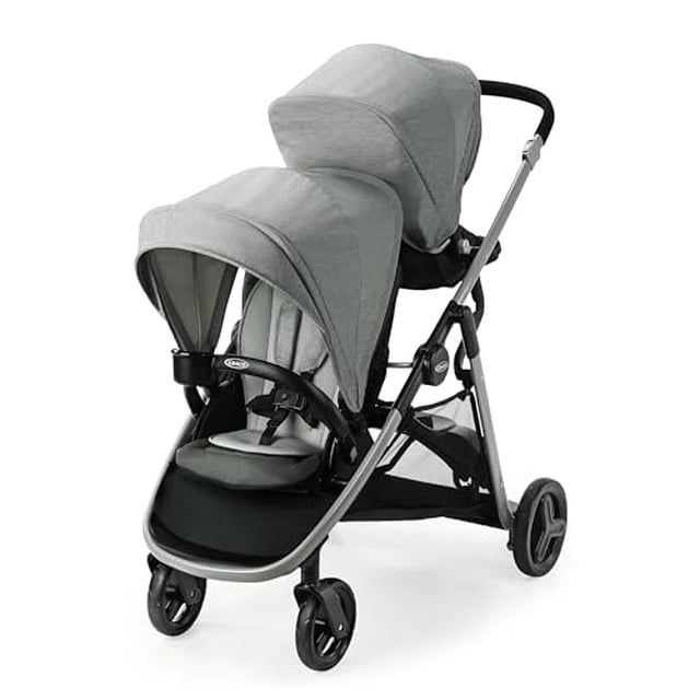 graco ready2grow lx 2.0 double stroller features bench seat and standing platform options, clark — Carriola doble Graco Ready2Grow LX 2.0 en gris clark, vista 3/4 frontal con capotas grandes; chasis metálico negro, dos asientos acolchados (superior e inferior), barra frontal desmontable con portavasos, gran canasta inferior y ruedas delanteras giratorias. Se pliega como carriola individual y el asiento trasero se convierte en banco o plataforma de pie, ideal para gemelos o hermanos de edades cercanas.