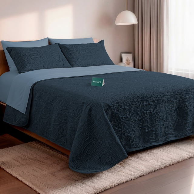 set de 7 piezas edredón ligero y sábanas matrimonial dreamlab, colcha para cualquier temporada, hipoalergénica, diseño moneda azul noche, azul mezclilla — Vista frontal centrada de cama matrimonial con colcha ligera azul noche (210 x 240 cm) de microfibra doble cepillado con patrón moneda y relieve texturizado; sábana plana y fundas en tono azul mezclilla más claro; incluye 2 fundas decorativas (71 x 48 cm) y 2 fundas de almohada estándar (51 x 76 cm); ambiente de dormitorio con lámpara colgante y cortina clara. Apto también como colcha ligera para sofá o sala pequeña.