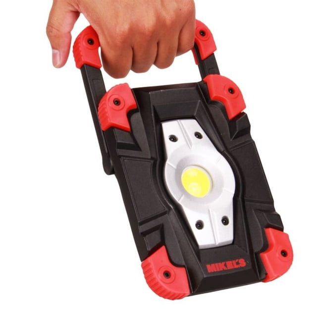 lámpara reflector led (10w recargable) uso rudo negro Mikel´s, vista frontal en 3/4 sostenida por una mano con asa plegable. Carcasa rectangular negra con refuerzos rojos en las esquinas, lente circular LED central sobre placa metálica plateada; para iluminación automotriz y taller, también apta para trabajos y señalización temporal.