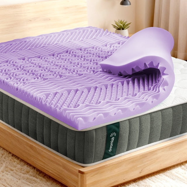 colchoneta de memory foam dreamlab con infusión de lavanda topper de 5cm matrimonial Lavanda, vista 3/4 frontal sobre colchón matrimonial. Topper de 5 cm de espuma viscoelástica texturizada con zonas contorneadas y perfiles ondulados que muestran alivio de puntos de presión; esquina levantada revela grosor; etiqueta Dreamlab visible. Ideal para añadir suavidad a colchones firmes y reducir transferencia de movimiento.