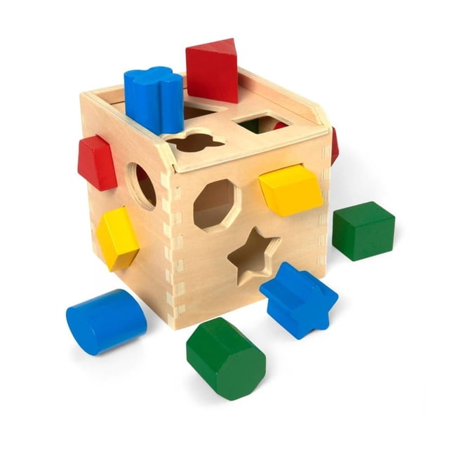 cubo de madera Melissa & Doug spinmaster para clasificar formas, vista frontal en 3/4 con tapa; cubo natural de madera dura con orificios recortados (estrella, hexágono, corazón, círculo, cuadrado, triángulo) y 12 bloques grandes multicolores (rojo, azul, verde, amarillo) alrededor y encima, piezas hacen ruido al caer y fomentan reconocimiento de formas y colores, también apto para juegos de clasificación en viajes.