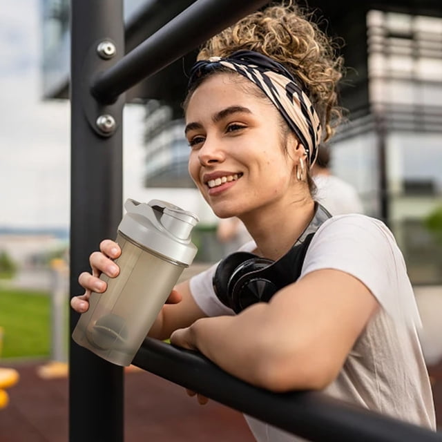 botella de agua deportiva portátil 500ml | taza para shaker de proteínas con báscula para mezclar, tapa abatible a prueba de fugas, agitador incorporado, boca grande fácil de limpiar | gimnasio muestra un shaker RINYA gris de 500 ml en vista 3/4 frontal con tapa abatible cerrada y boquilla visible, agitador integrado en la base, cuerpo cilíndrico ligero con agarre de una mano; fondo de parque y mujer apoyada en barra durante entrenamiento; también suele usarse para llevar polvos y mezclas en salidas de fitness.