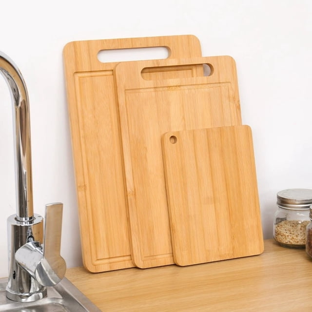 tabla de picar de doble cara | tabla de cortar antideslizante grande para cocina, conjunto de tres tablas tipo bambú en tonos madera claro, vista frontal 3/4 con asas empotradas y canal perimetral para líquidos; una tabla pequeña con orificio para colgar, apoyadas en encimera junto a fregadero, aptas para carne, verduras y queso; también suelen usarse para servir tablas de quesos.