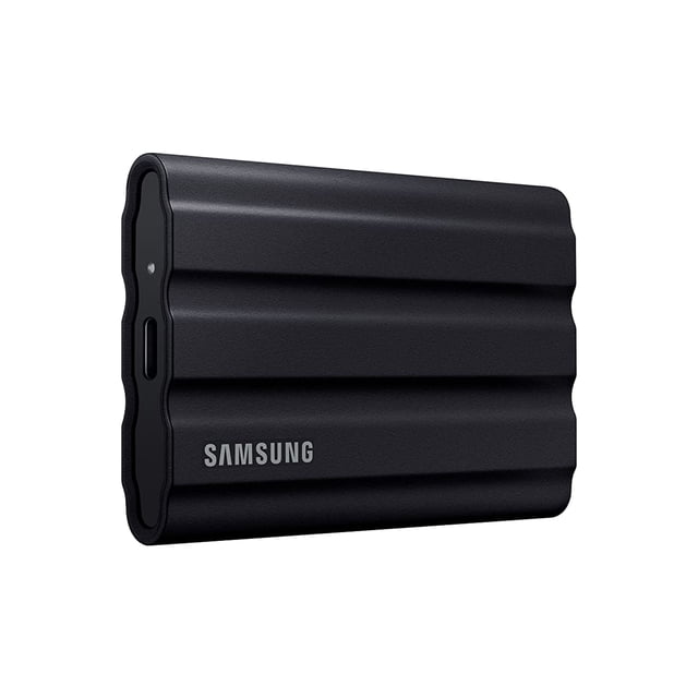 ssd portátil samsung t7 shield 2tb usb 3.2 gen2 negro, vista 3/4 frontal con carcasa negra texturizada y relieves ondulados, logotipo Samsung plateado, puerto USB-C y pequeño LED visibles; resistente según especificaciones IP65 y diseñado para transferencia rápida de archivos; también es adecuado como unidad externa portátil para respaldar sesiones de fotografía.