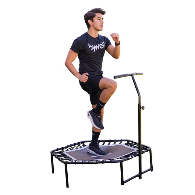 trampolin para aeróbicos uso rudo bywell fitness yoga — trampolín hexagonal de 48 pulgadas con bastidor metálico negro, superficie de salto gris y ligas elásticas visibles; vista 3/4 con persona haciendo step sujetada a manija ajustable; pensado para rutinas de aeróbicos y yoga en interiores y también suele usarse para ejercicios de estabilidad y cardio ligero.
