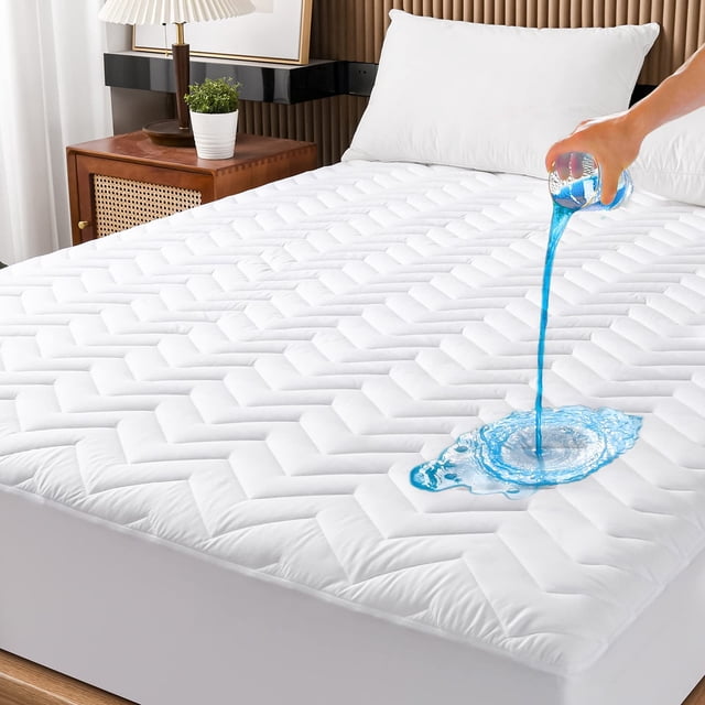 protector de colchón impermeable unilibra king size blanco protector King acolchado en patrón geométrico, vista 3/4 frontal con faldón elástico tipo funda bajera que cubre los laterales; mano vertiendo líquido azul sobre la superficie para mostrar la función 100% impermeable y la apariencia transpirable; habitación con mesita y almohada blanca; suele usarse también en camas de invitados.