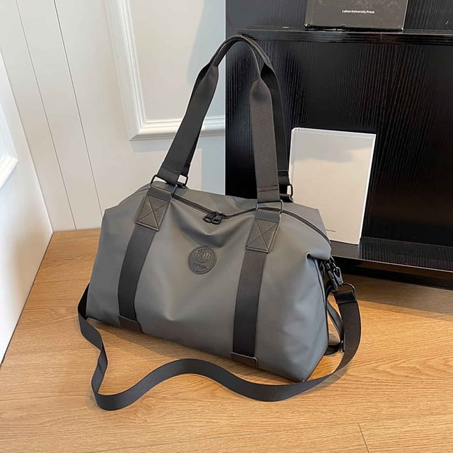 maleta cruzada plegable de gran capacidad • organizador multicompartimento • separación húmeda/seca • equipaje de mano Bolsa RINYA gris multicolor en vista frontal 3/4, bandolera plegable con asas dobles y correa ajustable desmontable; tela resistente, cierre superior de cremallera y logotipo circular frontal; diseño ancho con bolsillos laterales y compartimento dedicado húmedo/seco, suficiente para 3–5 días de ropa; apoyada sobre piso de madera junto a mueble, útil como bolsa extra de viaje o para entrenamiento.