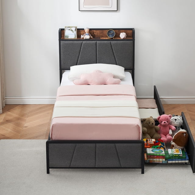 yeshouse cama individual con usb y 2 cajones, tapizada en lino, sin base, estructura silenciosa, gris — vista frontal centrada de cama individual tapizada en lino gris con cabecera acolchada de paneles y repisa superior con puerto USB integrado y ranuras para gestión de cables; ropa de cama rosa claro y cojín en forma de nube; dos cajones grandes a la derecha, uno abierto, mostrando juguetes y cajas sobre rieles de bolas; estructura metálica baja con patas antideslizantes y espacio bajo la cama visible (10.6"). Ahorro de espacio y almacenamiento oculto para habitación infantil o de invitados.