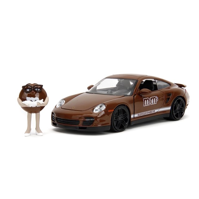 coche de juguete Jada Toys M&M's 1:24 Porsche 911 Turbo fundido a presión con figura — Vista 3/4 frontal de un Porsche 911 Turbo 2007 marrón a escala 1:24 con llantas negras de cinco radios, neumáticos de goma premium, calcomanía M&M's en la puerta y franjas laterales blancas; incluye figura marrón fundida a presión de 2.75 pulgadas con gafas. Vehículo con capó, puertas y maletero abatibles y ruedas de trabajo, adecuado para coleccionistas y juego a partir de 13 años; a menudo usado también como pieza de vitrina para fanáticos de autos en miniatura.
