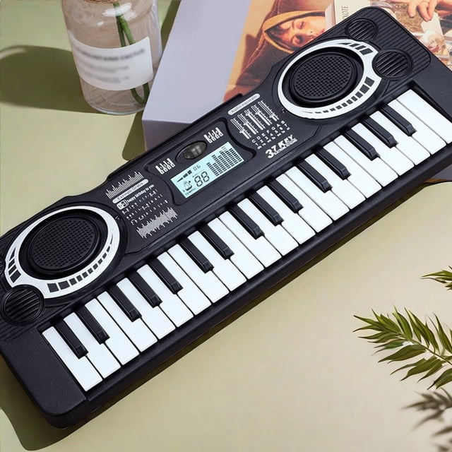 teclado electrónico piano 37 teclas | instrumento musical digital multifunción | órgano electrónico portátil para principiantes con modos de canto y piano | compacto 13,78 x 5,11 pulgadas | funciona — 3/4 frontal de un teclado negro de 37 teclas con dos altavoces integrados, pantalla LCD central y botones de ritmo/volumen, carcasa de plástico compacto (35 x 13 cm) y compartimento para 3 pilas AA; fondo con caja y planta, ideal como teclado educativo para niños y también adecuado para uso en clase o viajes cortos.